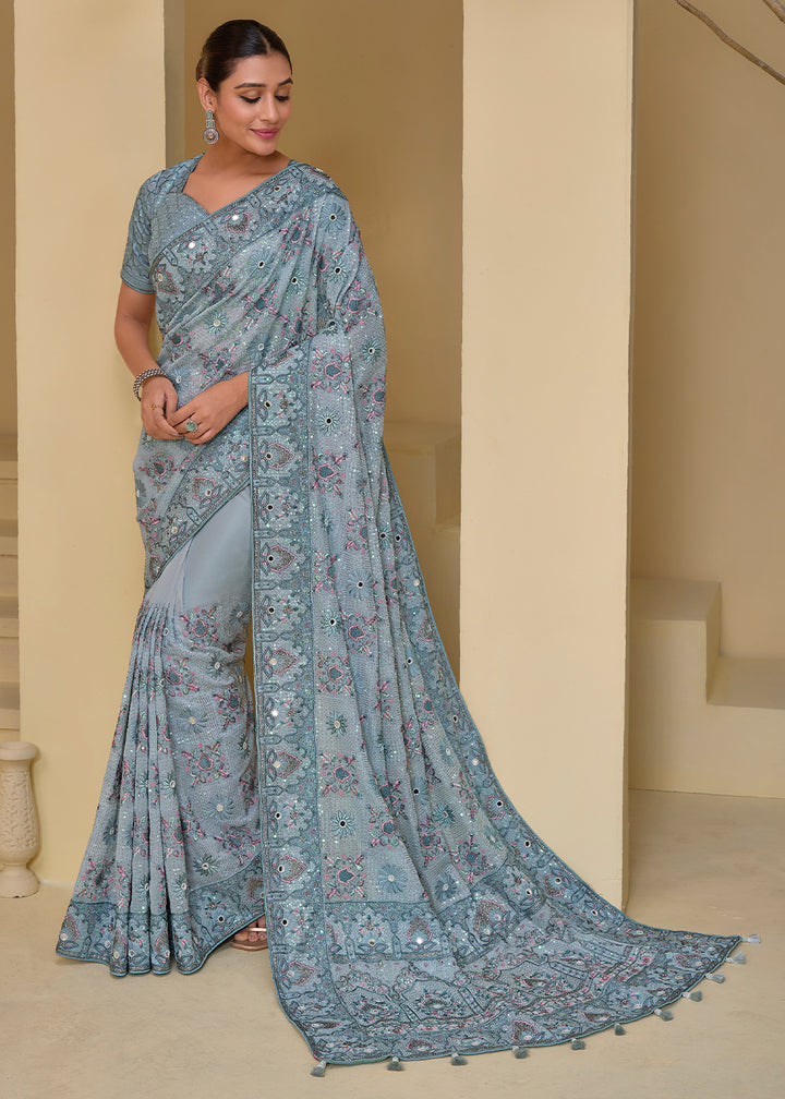 A woman wearing a bluish-grey Banarasi Tuscany silk saree with floral and geometric embellishments, posing in a classical stance with one hand on her hip.