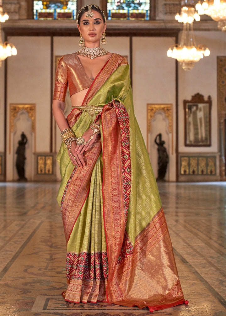 A woman showcasing a light olive green Banarasi silk saree with rich patola design and zari highlights, along with a red and gold brocade silk blouse piece.